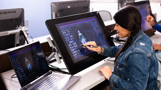 A smiling student sits at a table in a classroom and uses digital modeling software to create a 3D character.