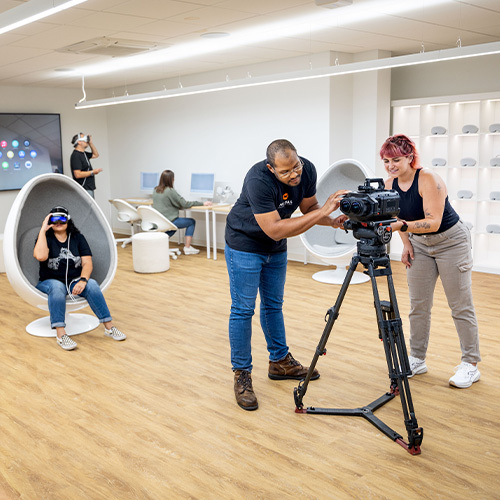 Students and instructors in a technology classroom work with specialty VR cameras, use VR headsets, and work on desktop computers.