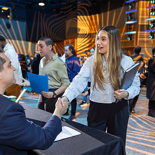 A student at a career fair smiles and shakes hands with an employer. Other students and employers mill around the room and network.