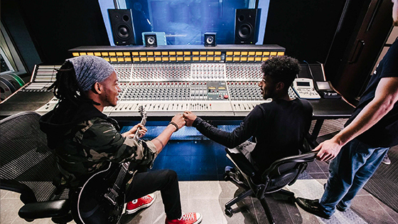 A student sitting with a guitar bumps fists with a student sitting at a sound board while an instructor looks on.
