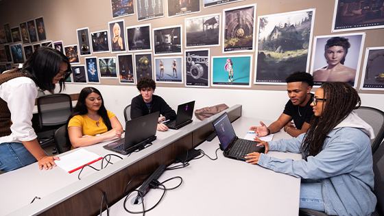 A group of students sit at tables in a classroom and work together on laptops. Printouts of digital designs are on the wall behind them.