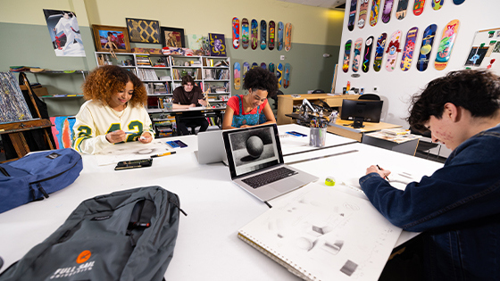 Students sit at tables in a classroom with colorful prints and skateboards on the wall. They are drawing by hand and using design editing software on laptops.