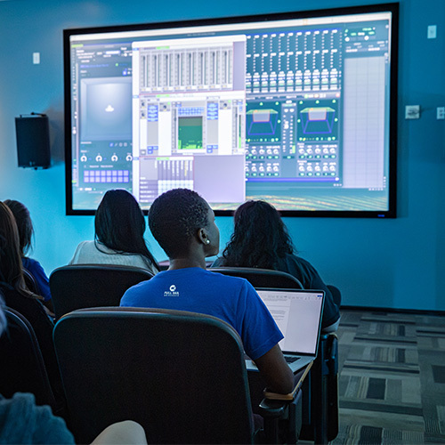 A group of students sit in chairs in a classroom and face a large digital display featuring Dolby Atmos software.