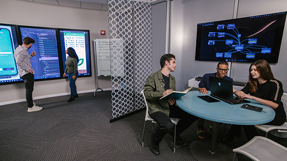 A group of students sit at a table in a classroom and work at a laptop. Two more students stand in front of large screens displaying coding software.