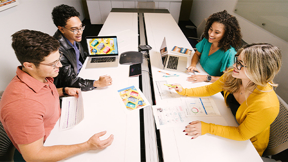 Four students sit and talk at a table in a small conference room. Laptops and printouts of bright designs and graphs are on the table.