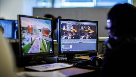 A student sits at a desk with dual monitors and uses video editing software on scenes from a film.