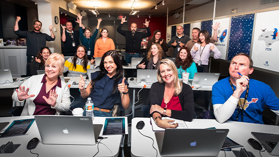 A large group of instructors sit at tables in a conference room. They are smiling and giving thumbs-up to the camera.