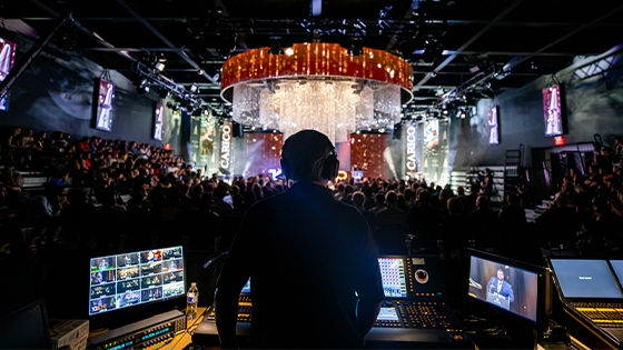An engineer works with sound and video equipment at the back of an event space. A large audience face the stage.