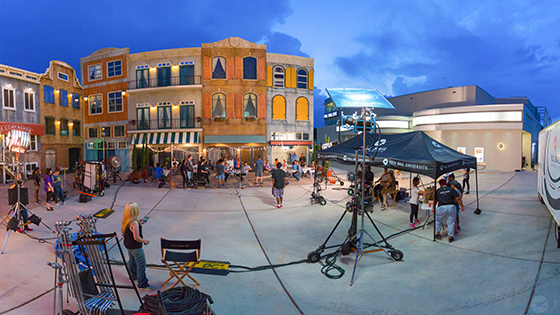 A large group of students work at cameras, lights, and sound equipment as they film a scene on Full Sail’s Backlot.