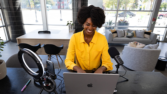 A smiling student in a bright yellow shirt sits at a desk with a laptop, microphone, and ring light.