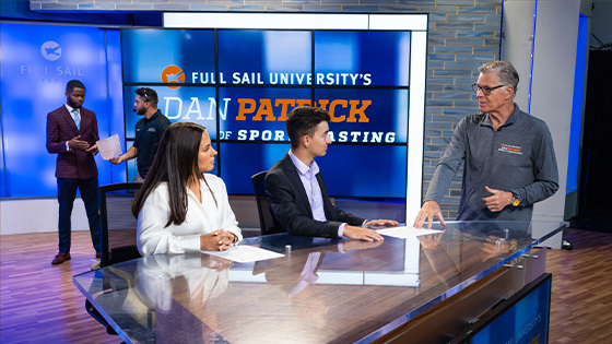 Two students sit at a news anchor desk in a TV studio and talk to Dan Patrick. The words Dan Patrick School of Sportscasting are on the wall behind them.