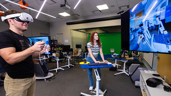A student using a VR headset and VR controllers stands next to a student typing on a keyboard. They are both looking at a screen displaying a VR room.